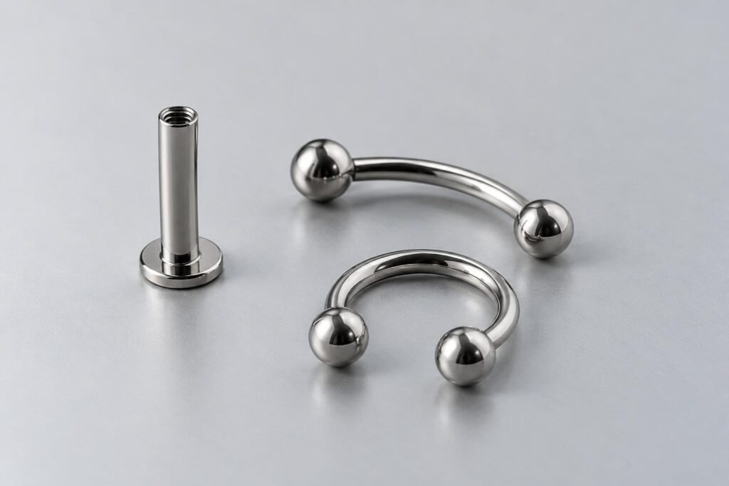 Gioielli da piercing dall’aspetto argentato: un labret con disco piatto, un barbell curvo e un anello circolare.