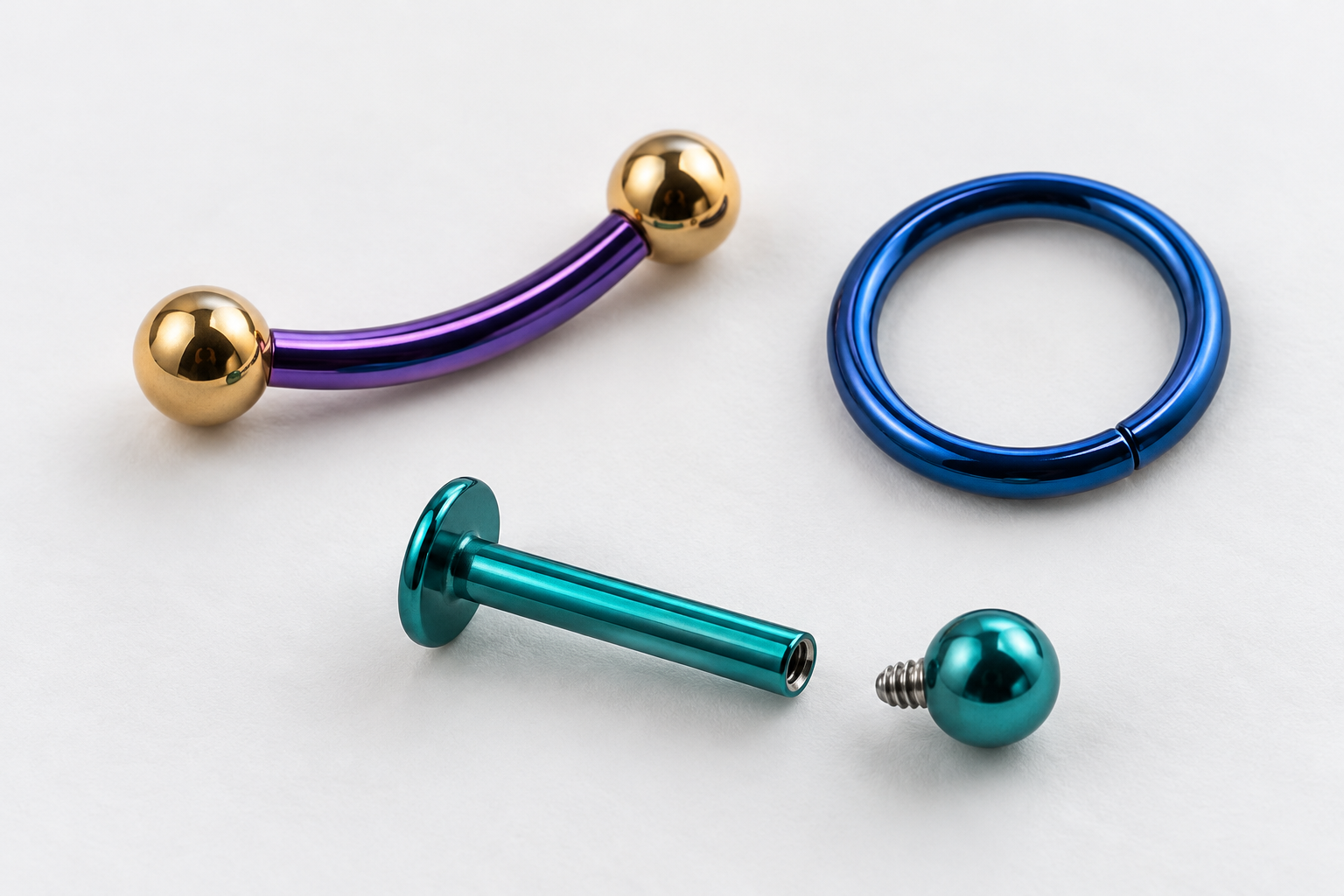 Tre gioielli da piercing su fondo bianco: un bilanciere curvo viola, un anello blu e un gioiello labret turchese con sfera separata.